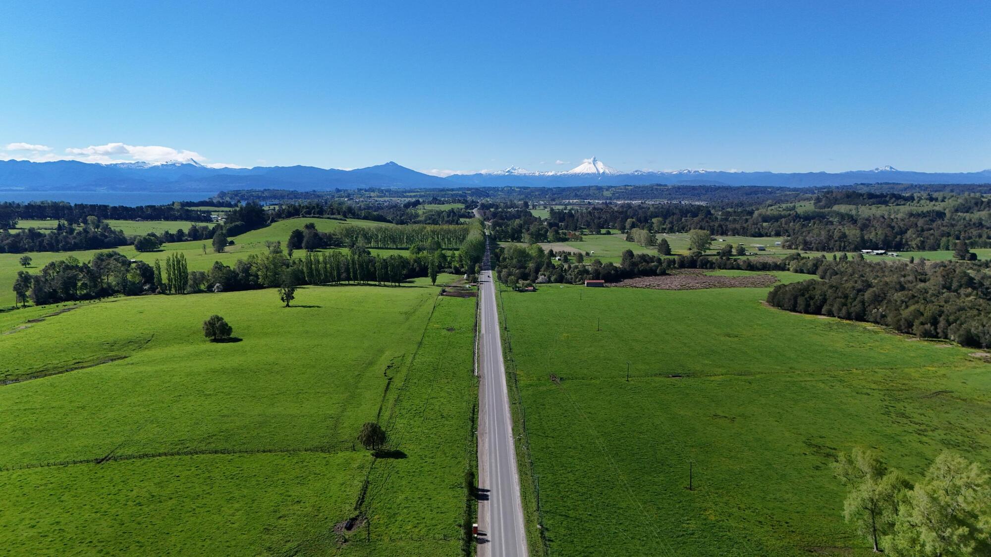 Vista aérea de una ruta rural recta entre praderas, con volcanes nevados y un lago al fondo